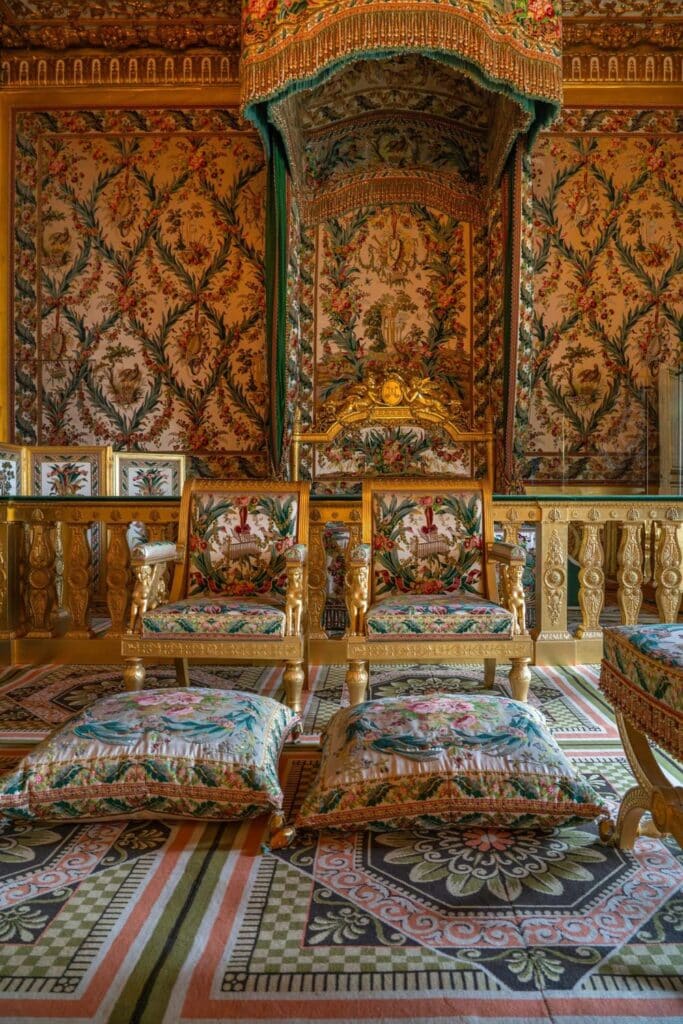Ornate royal bedroom in the Chateau de Fontainebleau featuring a gilded canopy bed, floral tapestry walls, and embroidered cushions on a patterned carpet—showcasing opulent French interior design.