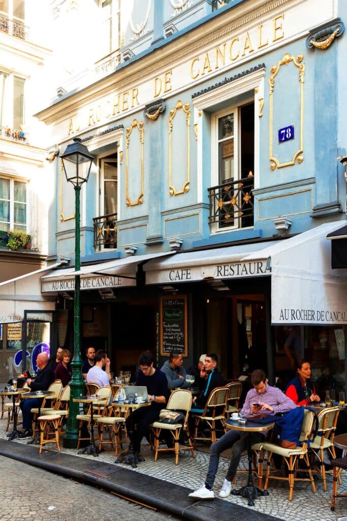 The blue and gold façade of Le Rocher de Cancale, a famous seafood restaurant on Rue Montorgueil. The outdoor terrace is filled with people enjoying oysters and French cuisine under a classic Parisian awning.
