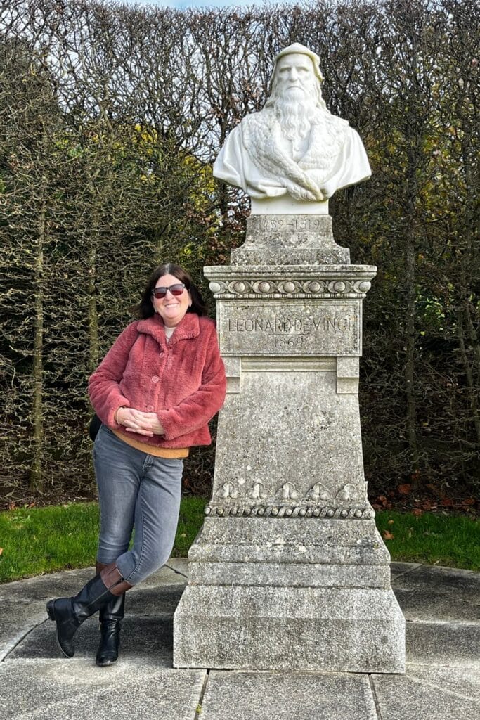 A stone bust of Leonardo da Vinci on a pedestal at Château d'Amboise, honoring the legendary artist and inventor. A visitor in a red coat poses beside it, framed by a hedge-lined garden.