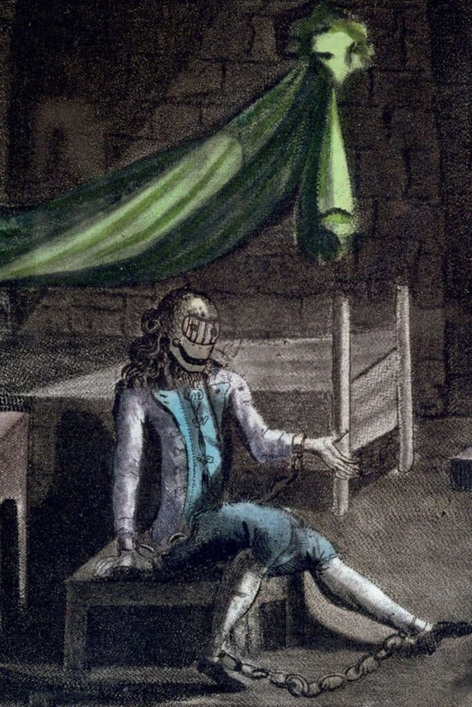 A haunting colored engraving of a prisoner in elegant clothing shackled to the wall of a stone cell, wearing the infamous iron mask. This fictionalized glimpse inside the Bastille evokes its mysterious and oppressive atmosphere.