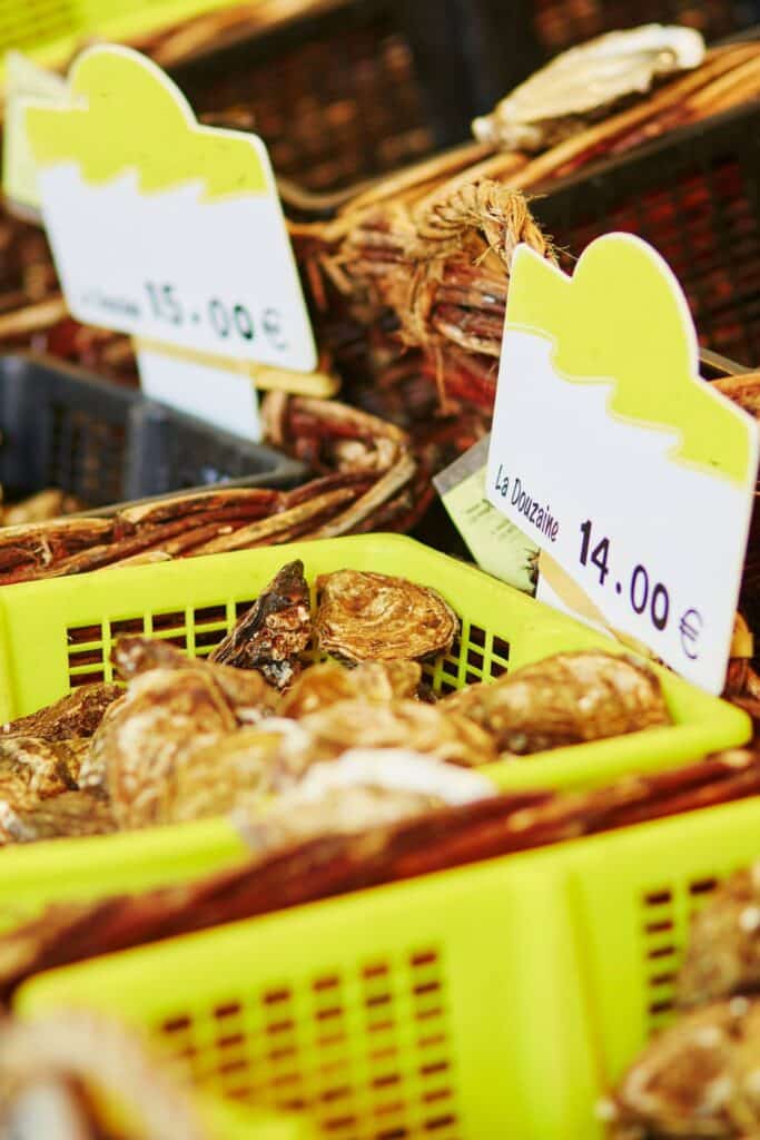 Fresh oysters displayed in bright yellow baskets at a seafood market on Rue Montorgueil. A price tag reading "La Douzaine 14,00€" emphasizes the affordability of this Parisian delicacy.