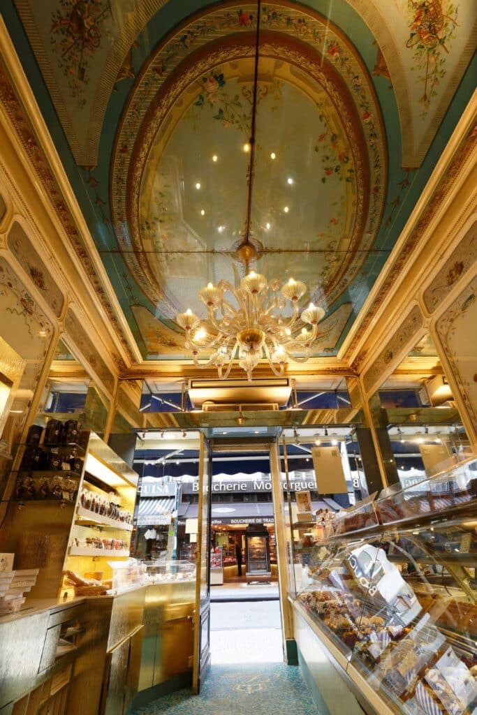 The opulent interior of Pâtissier Stohrer, one of the oldest pastry shops in Paris, located on Rue Montorgueil. A grand chandelier, ornate painted ceilings, and mirrored walls enhance the luxurious pastry display.