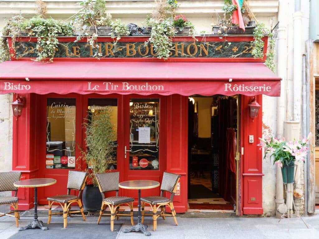 The vibrant red storefront of Le Tir Bouchon, a bistro and rotisserie on Rue Montorgueil. Outdoor seating with traditional woven bistro chairs and lush greenery above the awning add to its Parisian charm.