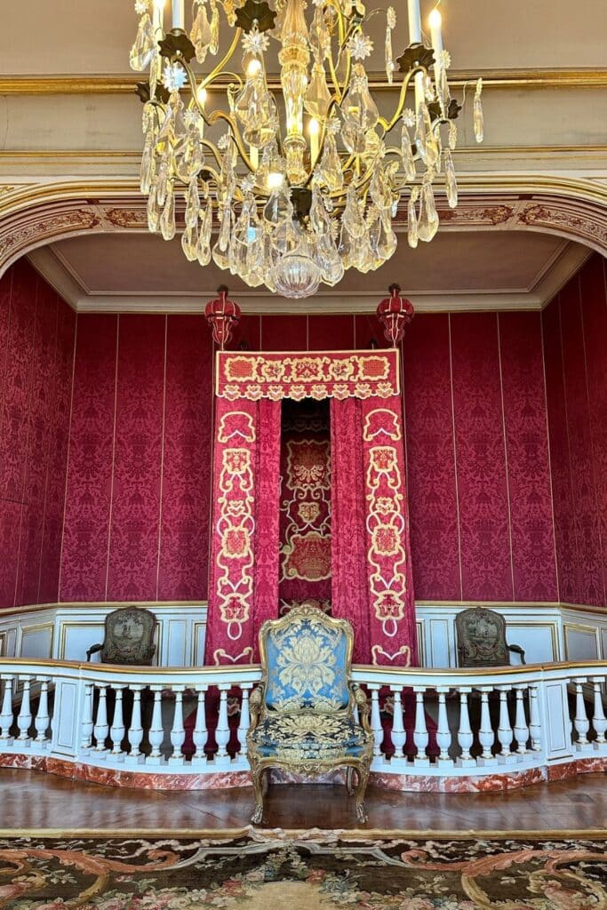 A lavish red and gold ceremonial space inside Château de Chambord, featuring a regal chair beneath an embroidered canopy, illuminated by a grand chandelier.