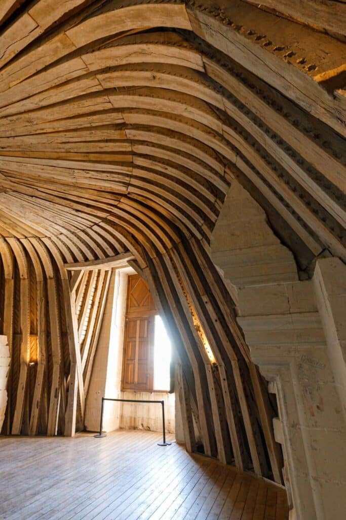 A rustic, vaulted attic space inside Château de Chambord, showcasing its aged wooden support beams curving towards an open window. The architectural details highlight the castle’s historic craftsmanship.