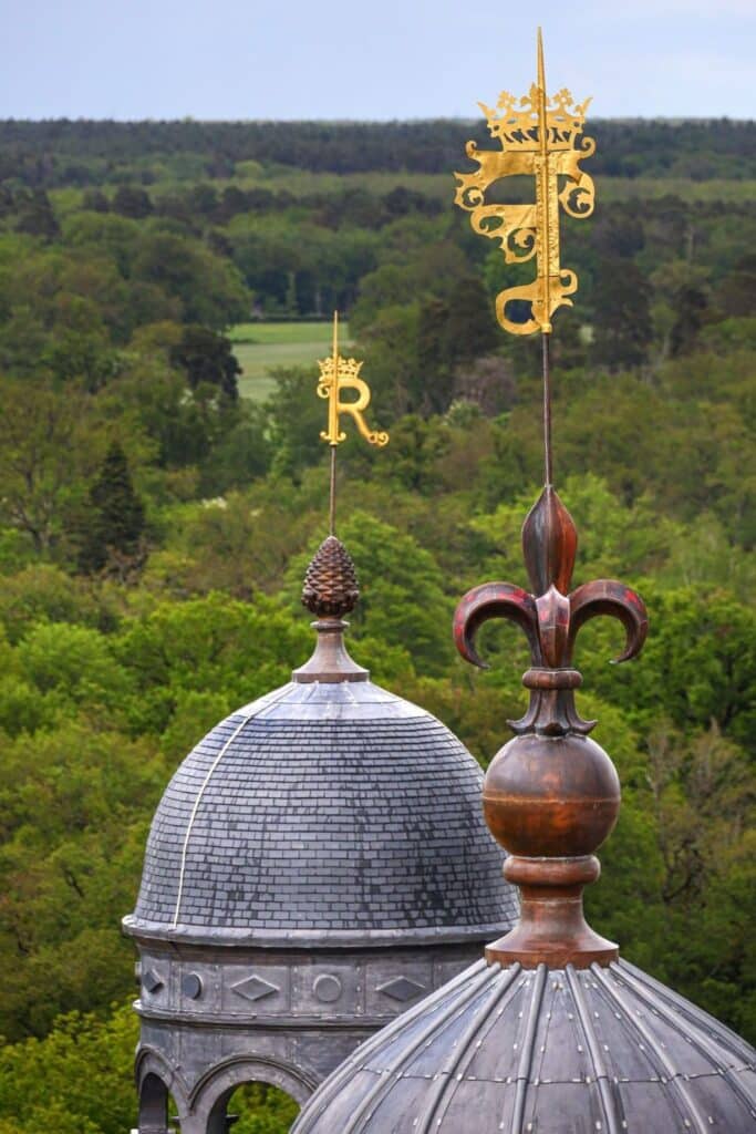 A close-up of ornate golden wind vanes atop Château de Chambord’s towers, featuring intricate Renaissance designs and crowned initials, set against the lush greenery of the Loire Valley.