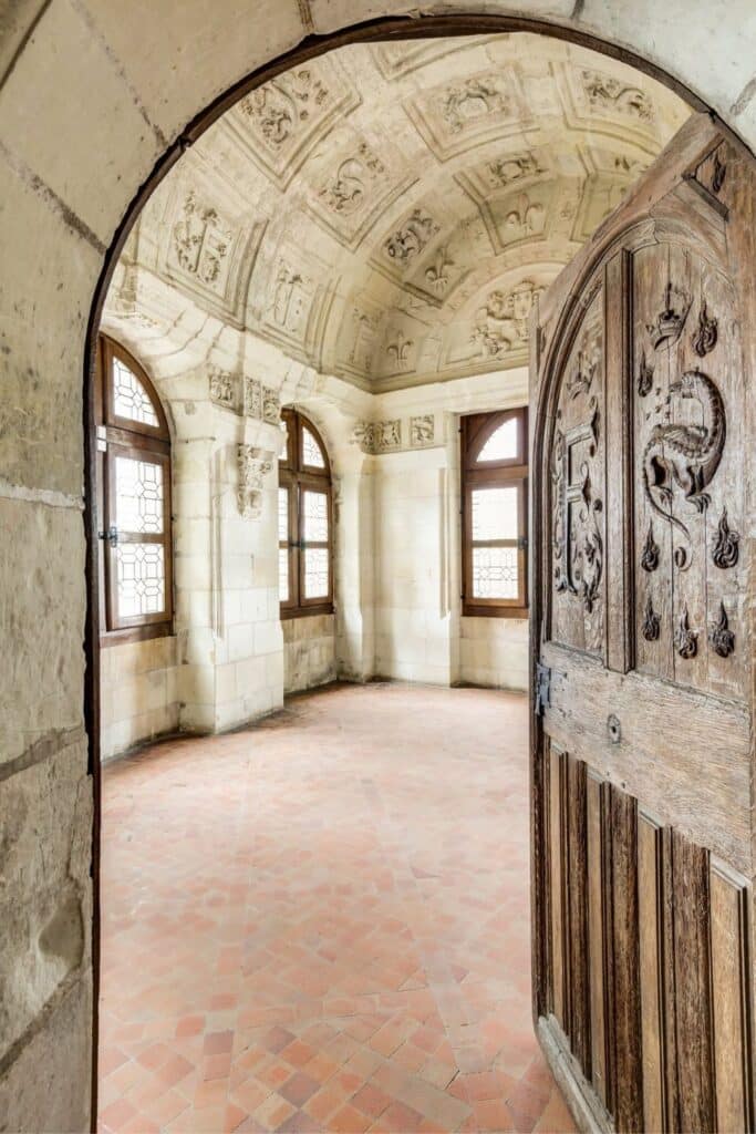 A grand stone chamber in Château de Chambord, viewed through an intricately carved wooden door. The ceiling is adorned with Renaissance reliefs featuring royal motifs.