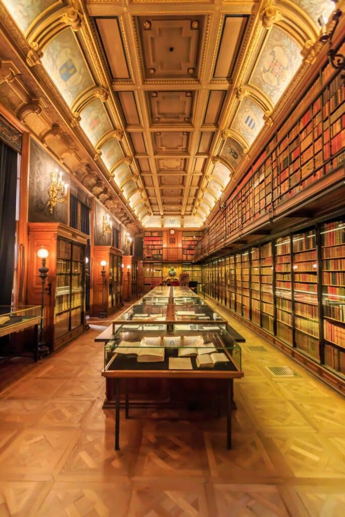 A majestic reading room in Château de Chantilly, illuminated by chandeliers, featuring a long table displaying antique books, with walls lined with elegant bookshelves.