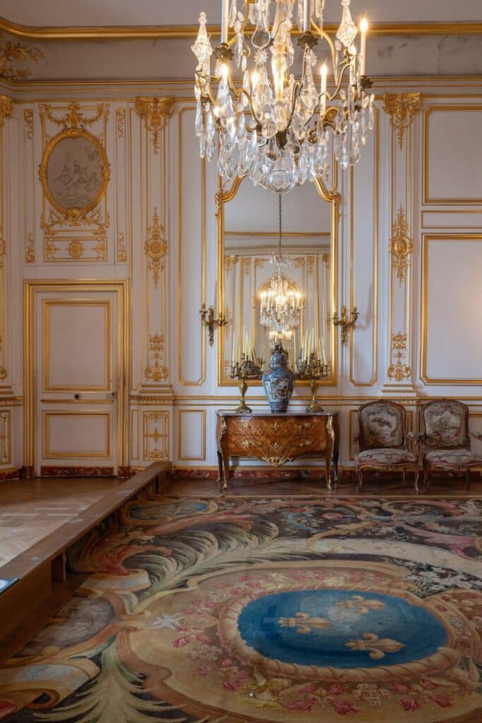 A luxurious interior in Château de Chambord featuring gold-trimmed walls, a sparkling chandelier, and a richly patterned carpet with royal fleur-de-lis motifs.