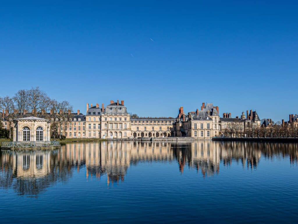 Panoramic view of the Chateau de Fontainebleau reflected on the calm waters of its grand pond, with a blue sky above and the pavilion standing prominently on the left.