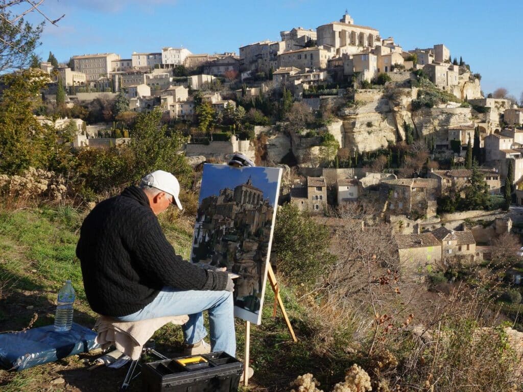 A man sits outdoors painting a canvas of the iconic hillside village of Gordes, capturing its stacked stone houses and sunlit charm—one of the most artistic things to do in Gordes.