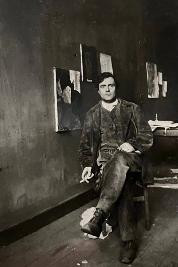 Black-and-white portrait of Amedeo Modigliani, a famous resident of Montmartre, seated in a sparse studio with abstract paintings on the wall behind him. He wears a rugged suit and holds a cigarette, embodying the bohemian spirit of the Parisian art scene.