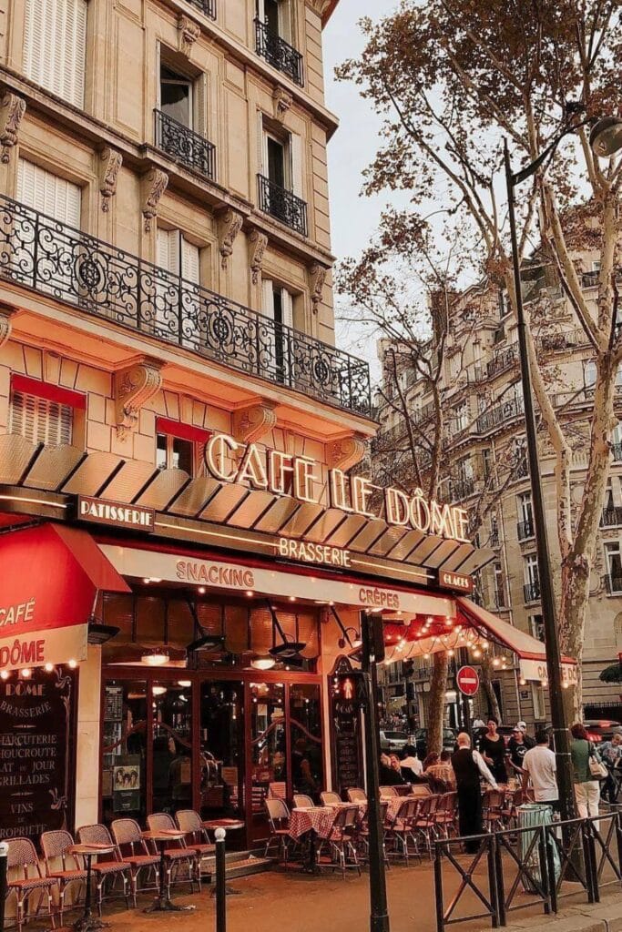 Charming street view of Café Le Dôme, a famous Paris cafe with classic red awnings, outdoor wicker seating, and golden signage glowing under soft lights on a Haussmann-style building corner.