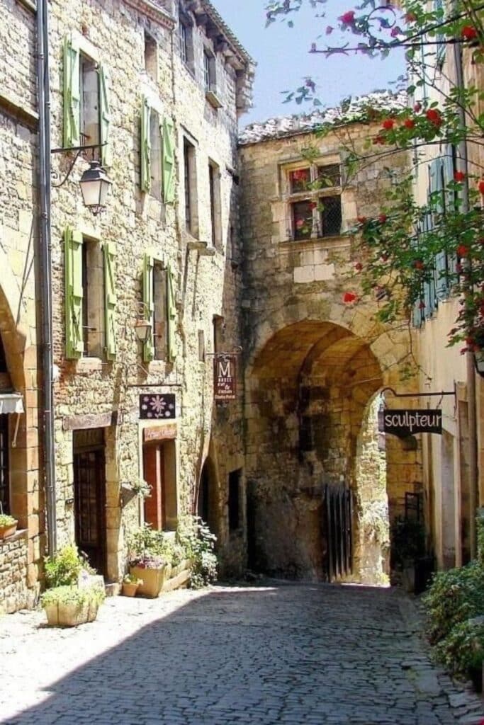 A narrow, stone-paved alley winds under an ancient archway in Cordes-sur-Ciel, framed by old stone houses with pastel shutters—classic ambiance of storybook towns in France.
