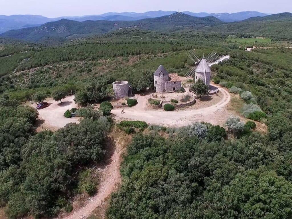 Aerial view of the historic windmills of Faugères nestled among rolling green hills, a tranquil countryside scene in the South of France's Languedoc region.