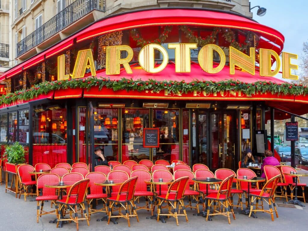 Bright red facade of La Rotonde, one of the most famous Paris cafes, adorned with festive greenery and iconic red-and-gold signage, with rows of red café chairs lining the sidewalk.