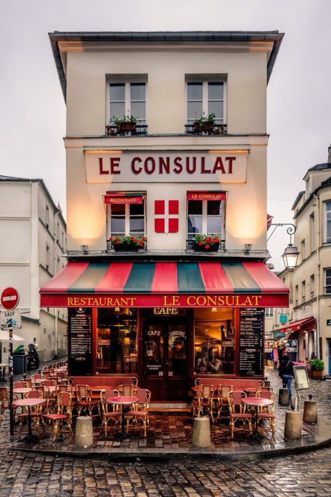 Rainy cobblestone street scene featuring Le Consulat, a famous Paris cafe in Montmartre with a striped red and green awning, vintage signage, and cozy bistro tables outside.