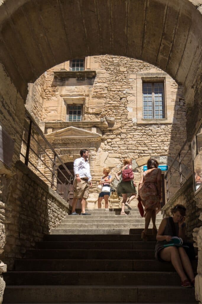 Visitors climb stone steps beneath an arched passageway in Gordes’ old village, surrounded by rustic walls and sunshine—an everyday slice of life among things to do in Gordes.