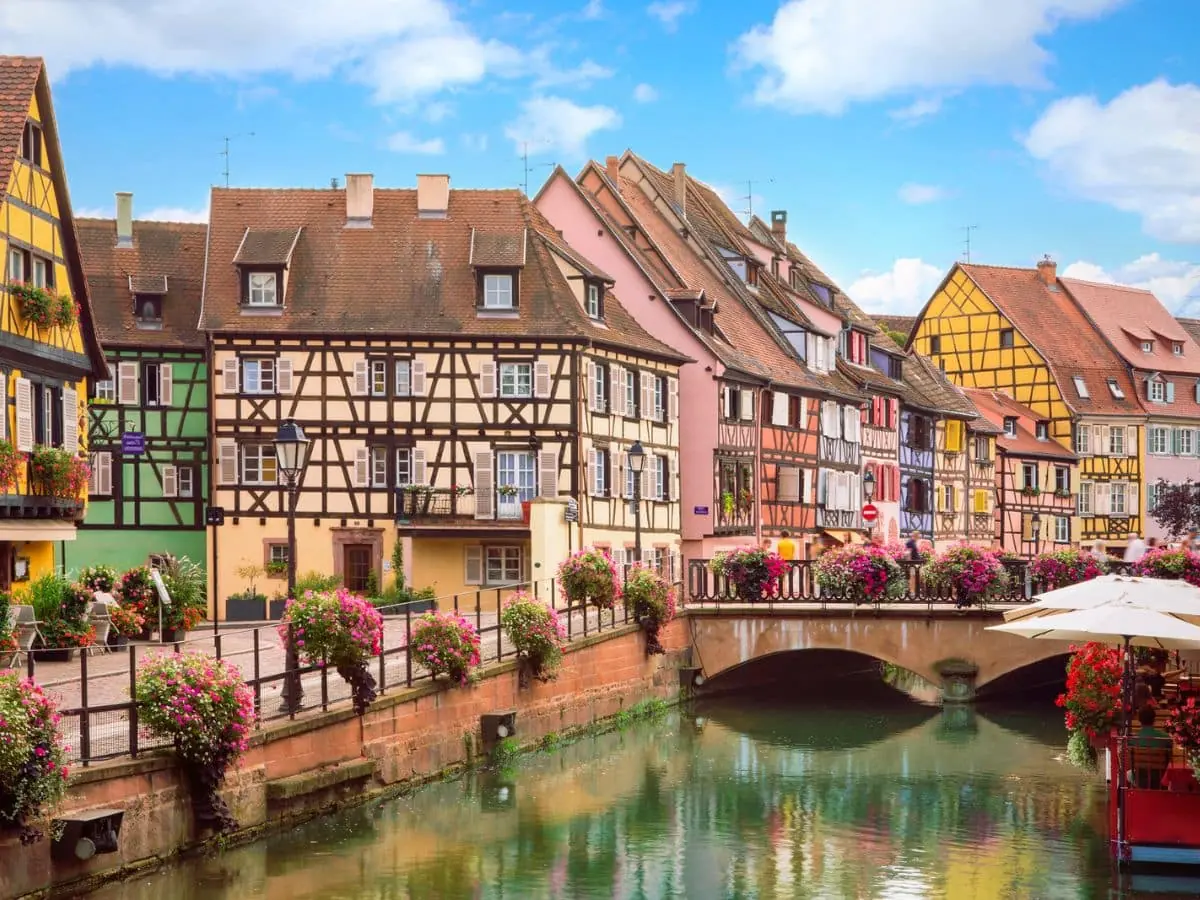 A scenic canal in Colmar lined with half-timbered, pastel-colored houses and vibrant flower arrangements, capturing the charm of the Alsace Wine Route.
