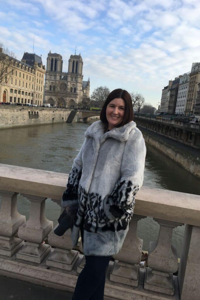 A woman in a light gray fur coat stands smiling on a stone bridge near Notre-Dame Cathedral, with the Seine River flowing behind her. This spot is one of the many scenic viewpoints showcasing historic bridges in Paris.
