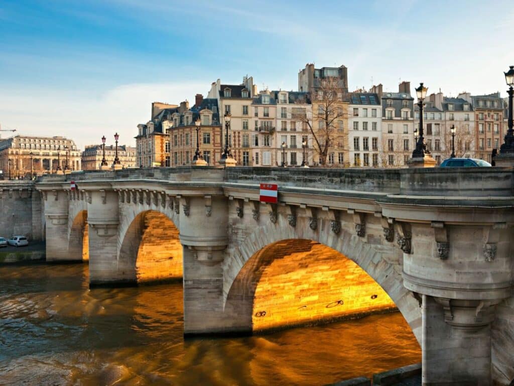 Pont Neuf, the oldest bridge in Paris, shines with golden light under its arches as the sun sets, illuminating the surrounding historic buildings.