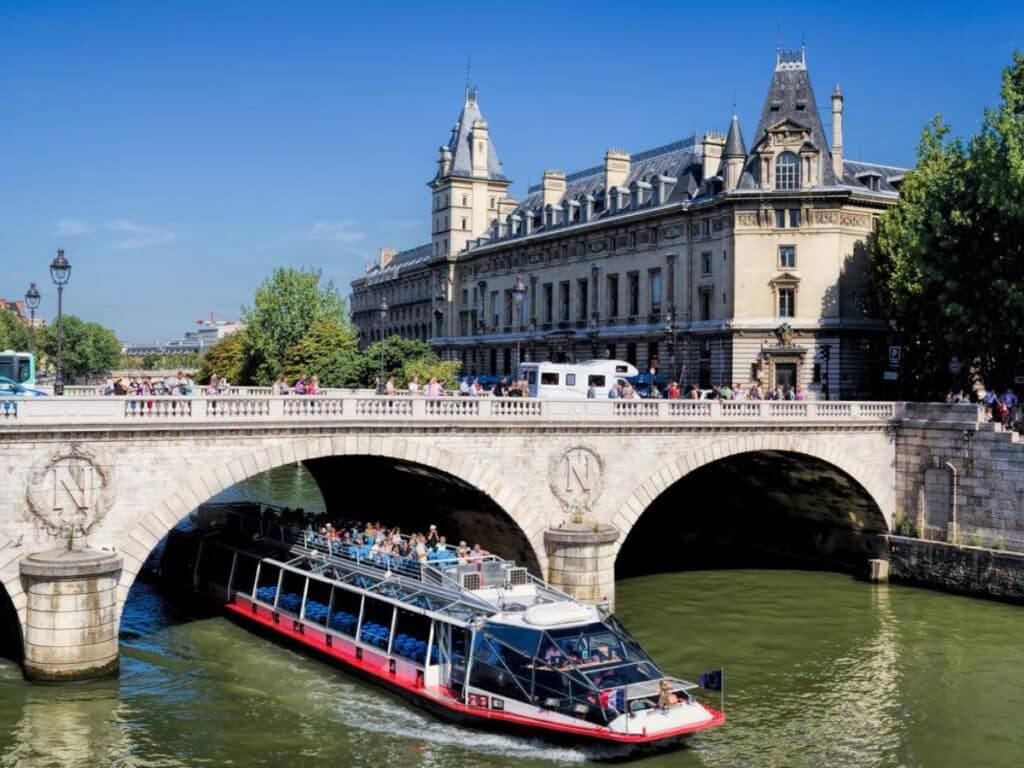 A riverboat cruise passes under Pont Saint Michel, one of the well-known bridges in Paris, with the Conciergerie towering beside and pedestrians enjoying views above.