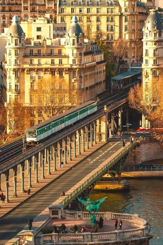 A metro train crosses Pont de Bir-Hakeim, an elevated bridge in Paris with pedestrian walkways below and classic buildings lining the Seine. This Paris bridge is known for its unique double-deck structure and cinematic views.