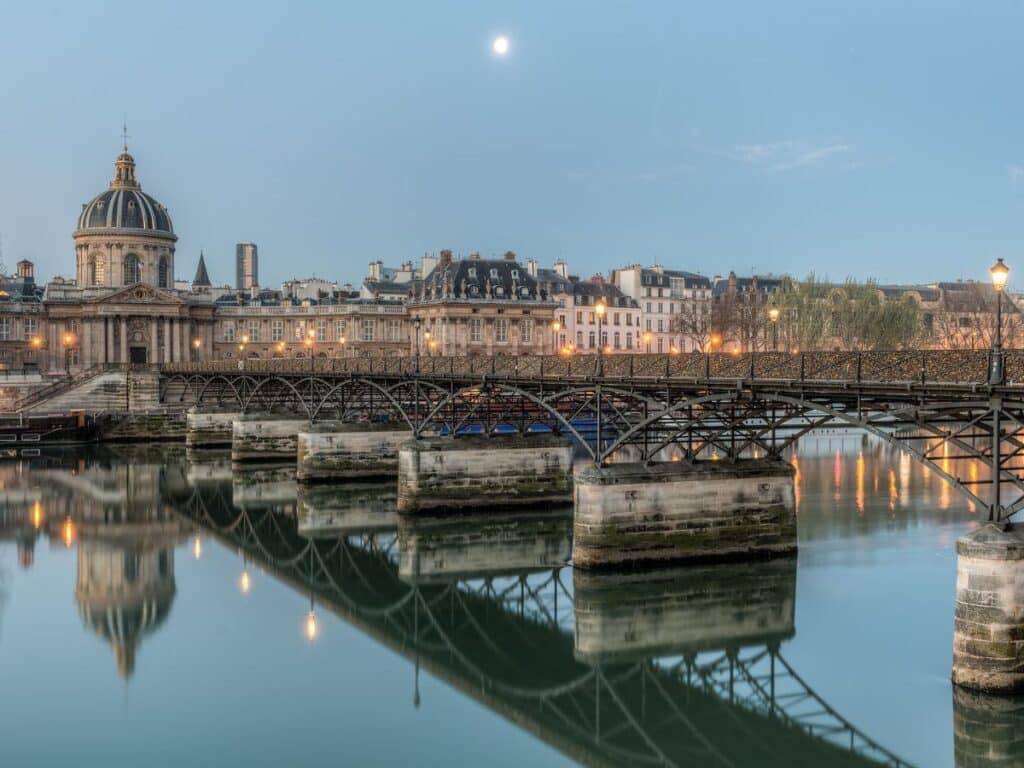 Pont des Arts glows softly in the evening light, reflecting in the calm Seine beneath the Institut de France and a full moon. This is one of the most romantic bridges in Paris, once famous for love locks.