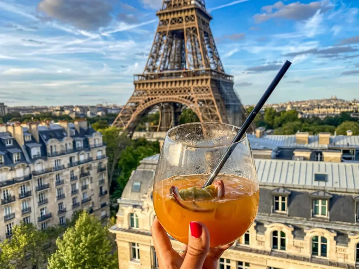 Hand holding a tropical cocktail in front of the Eiffel Tower with classic Haussmann rooftops in view, capturing a quintessential Paris rooftops moment.