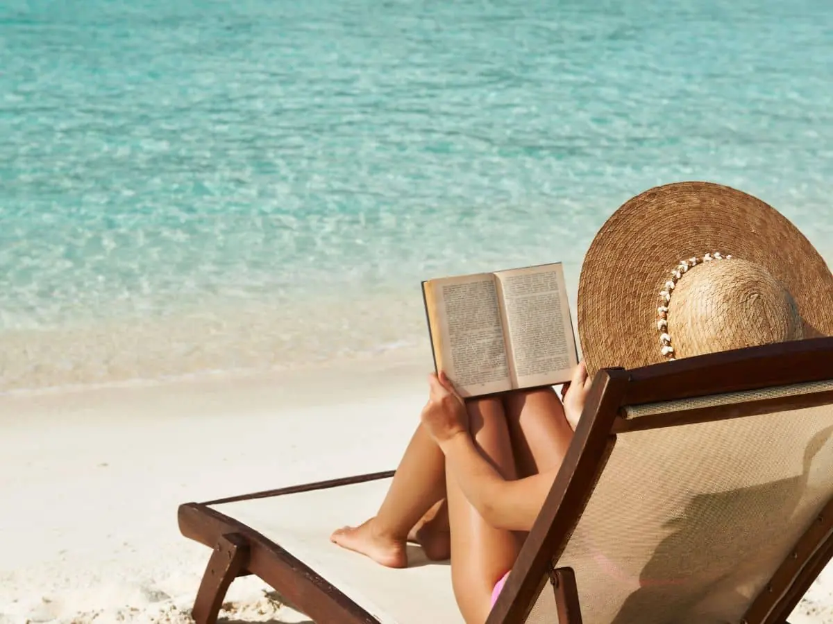 A woman in a sunhat lounging on a beach chair while reading a book beside turquoise waters—an idyllic scene that captures the vibe of enjoying summer beach reads set in France.