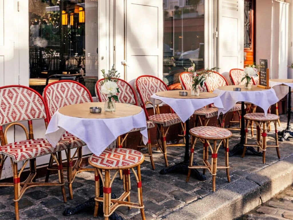 Parisian café terrace with red-and-white woven chairs and round bistro tables lined up along a cobblestone sidewalk, each table topped with a white cloth and a small flower vase — a classic setting reflecting slow-paced, proper café etiquette.