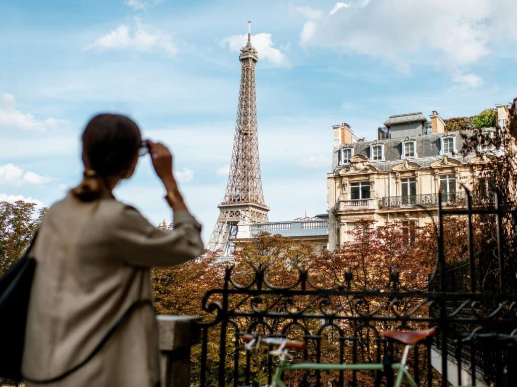 A woman in a neutral-toned coat looks toward the Eiffel Tower from a Parisian balcony, embracing Parisian chic with understated style and iconic city views.