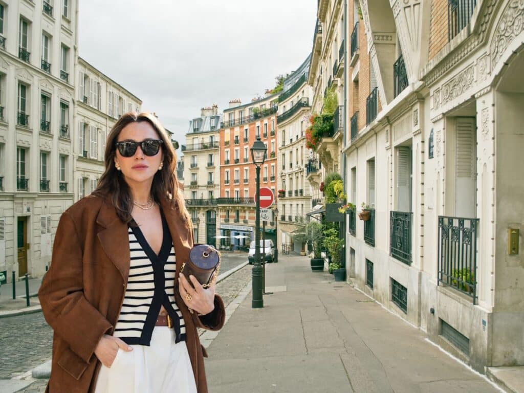 A woman stands confidently on a cobblestone street in Paris wearing an oversized brown coat over a striped top and wide white trousers, accessorized with dark sunglasses and a designer clutch. This look blends chic Parisian style with intentional layering, avoiding the fashion faux pas of skipping outerwear or structure.