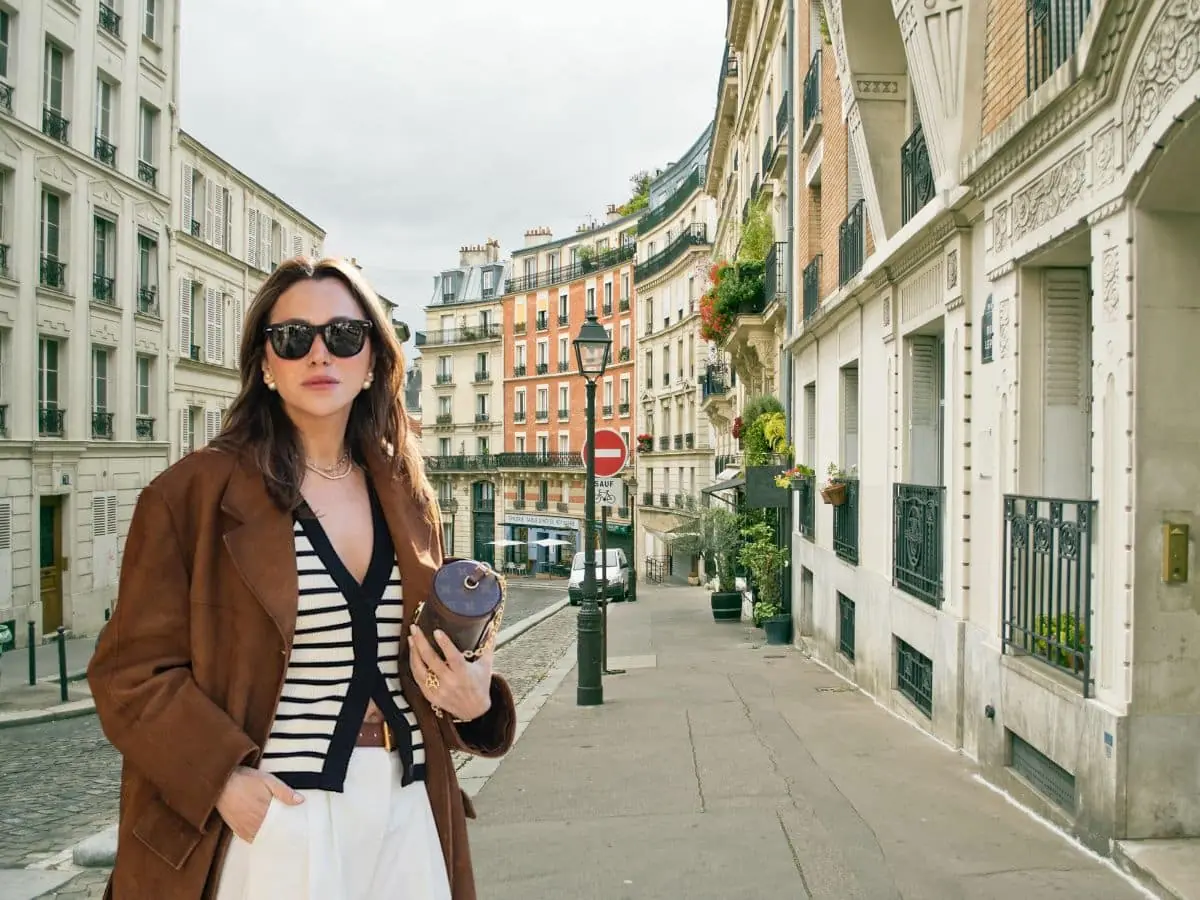 A woman stands confidently on a cobblestone street in Paris wearing an oversized brown coat over a striped top and wide white trousers, accessorized with dark sunglasses and a designer clutch. This look blends chic Parisian style with intentional layering, avoiding the fashion faux pas of skipping outerwear or structure.