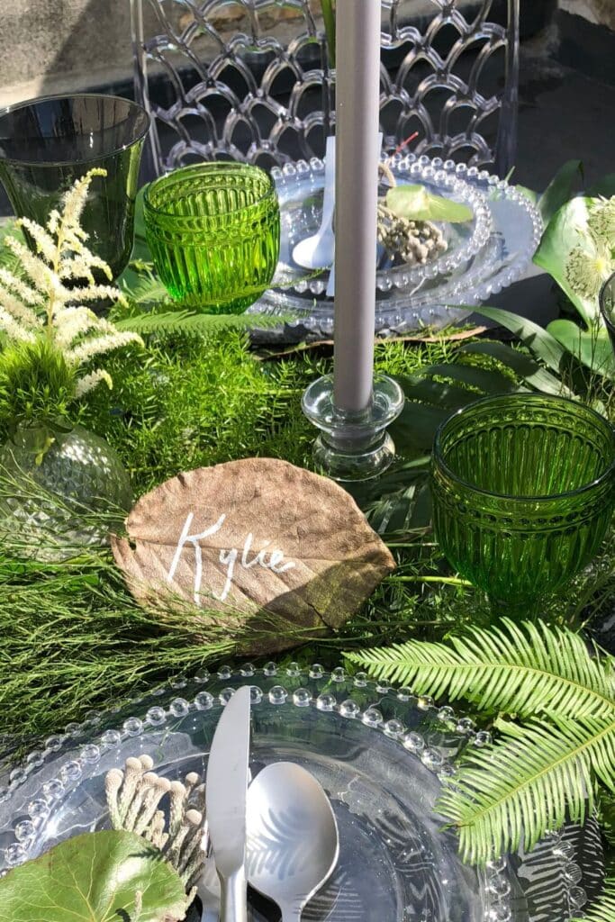 A beautifully arranged outdoor table setting with clear beaded glass plates, silverware, green glassware, and lush greenery. A dried leaf with the name "Kylie" in white script serves as a place card, adding a rustic personal touch.