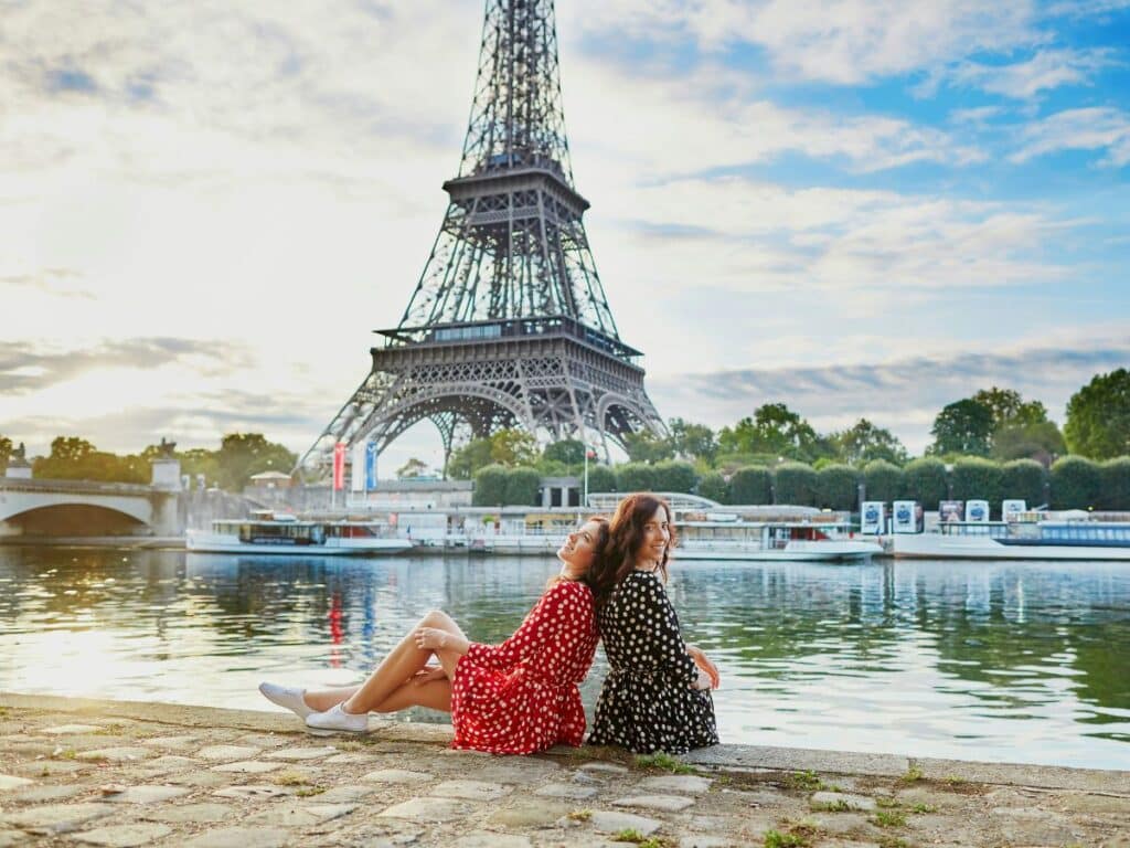 Two women sit by the Seine with the Eiffel Tower in the background, dressed in contrasting polka dot dresses—one in red, the other in black. Their effortlessly stylish looks embrace Parisian elegance and steer clear of flashy or overexposed clothing, a common fashion faux pas for tourists.