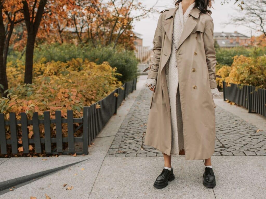 A woman wears a classic beige trench coat over a cozy knit outfit while walking through a park with autumn foliage—capturing the essence of Parisian chic in colder seasons.