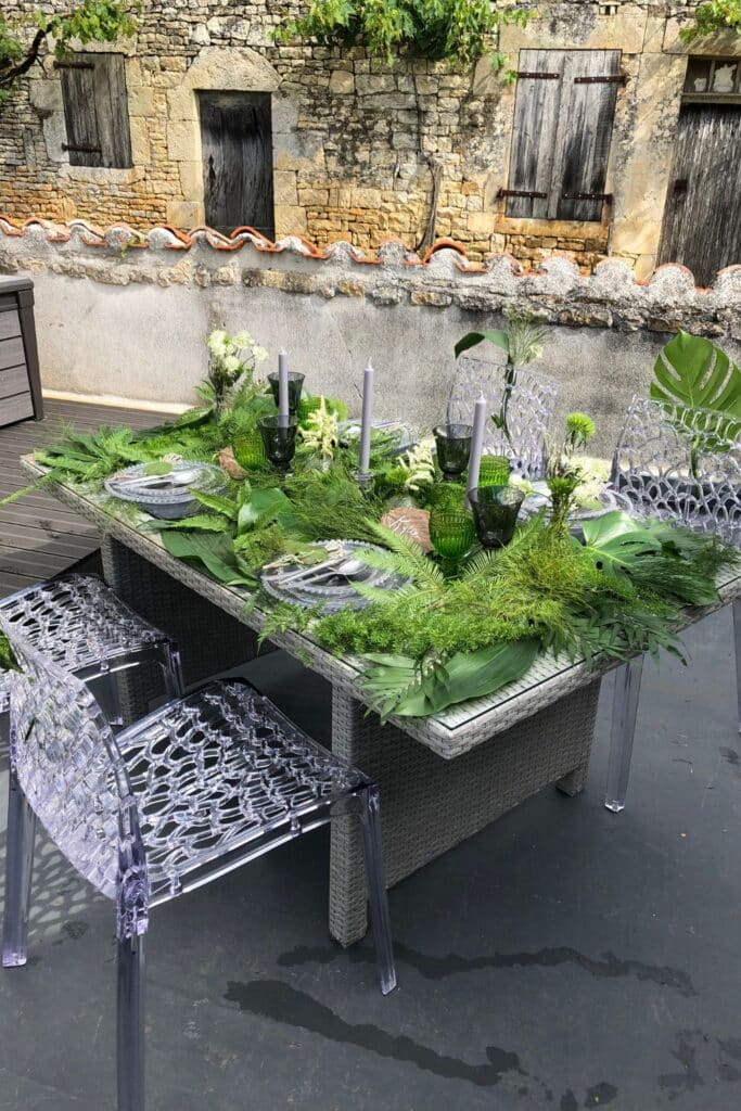 A nature-inspired summer garden tablescape set against a rustic stone wall. The table is adorned with vibrant greenery, candles, and green glassware, creating an inviting al fresco dining experience.