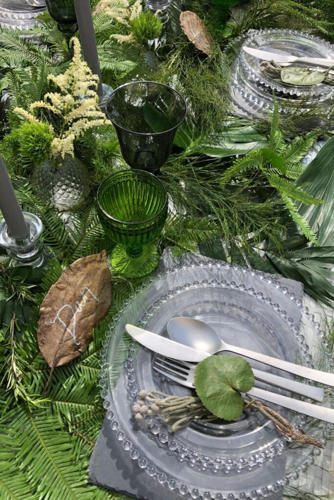 A close-up of a plate setting with a beaded glass plate, silver cutlery, and a dried leaf place card reading "Dan." The table is styled with fresh ferns, candles, and green accents for a lush springtime ambiance.