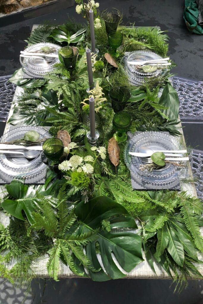 Overhead shot of a summer garden tablescape with large tropical leaves, ferns, and flowers cascading over the table. Beaded glass plates, green glassware, and personalized leaf place cards complete the look.