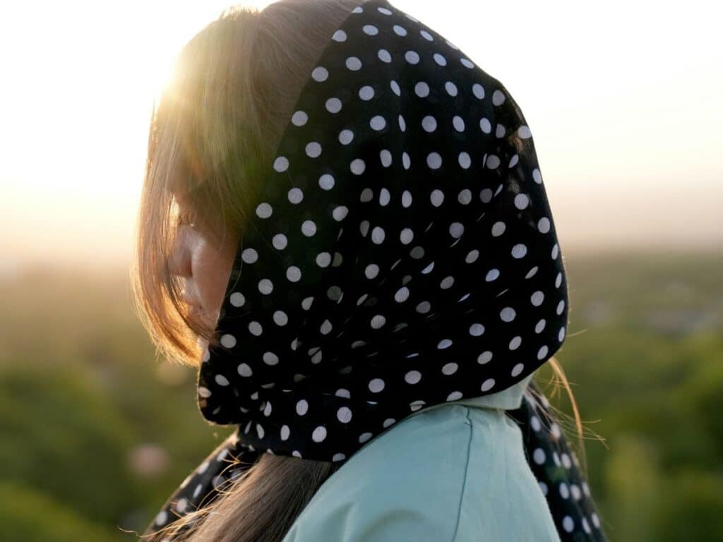 A woman stands in soft sunset light wearing a black scarf with white polka dots draped over her head, exuding timeless Parisian chic with effortless elegance.