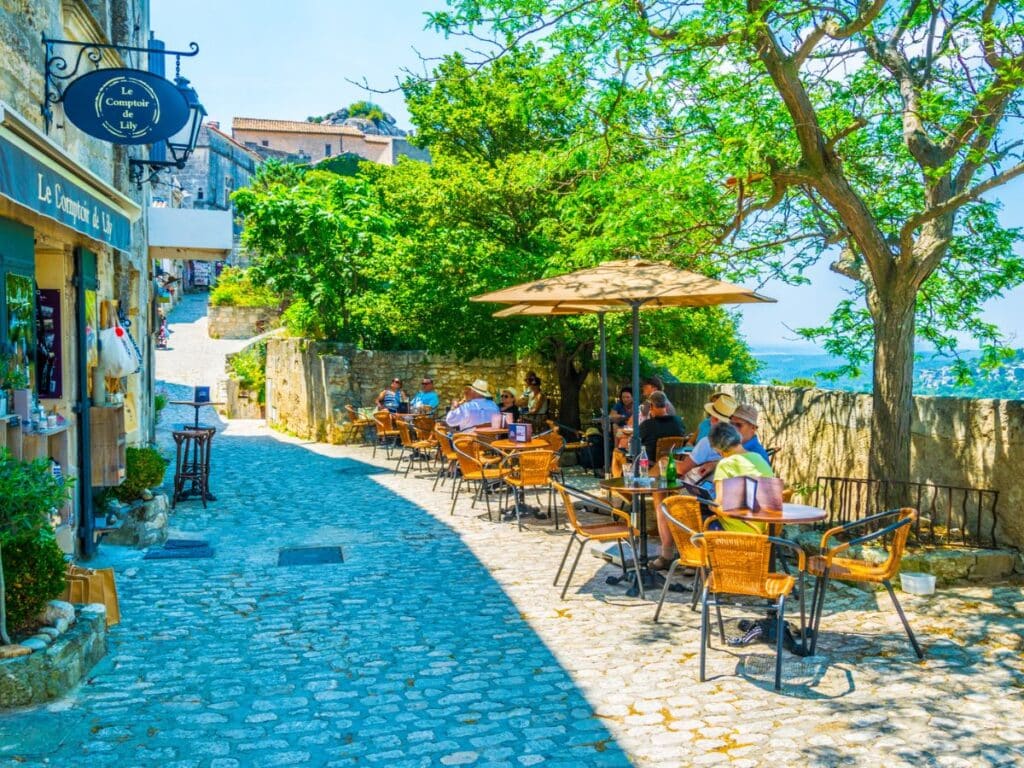 A cafe scene in Les Baux-de-Provence on a beautiful day with people sitting drinking under umbrellas next to stone wall.