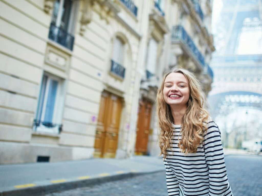 A smiling woman in a casual black-and-white striped long-sleeve shirt poses in front of the Eiffel Tower on a Parisian street. Her look is classic and comfortable without falling into the trap of overdone or inappropriate tourist fashion.