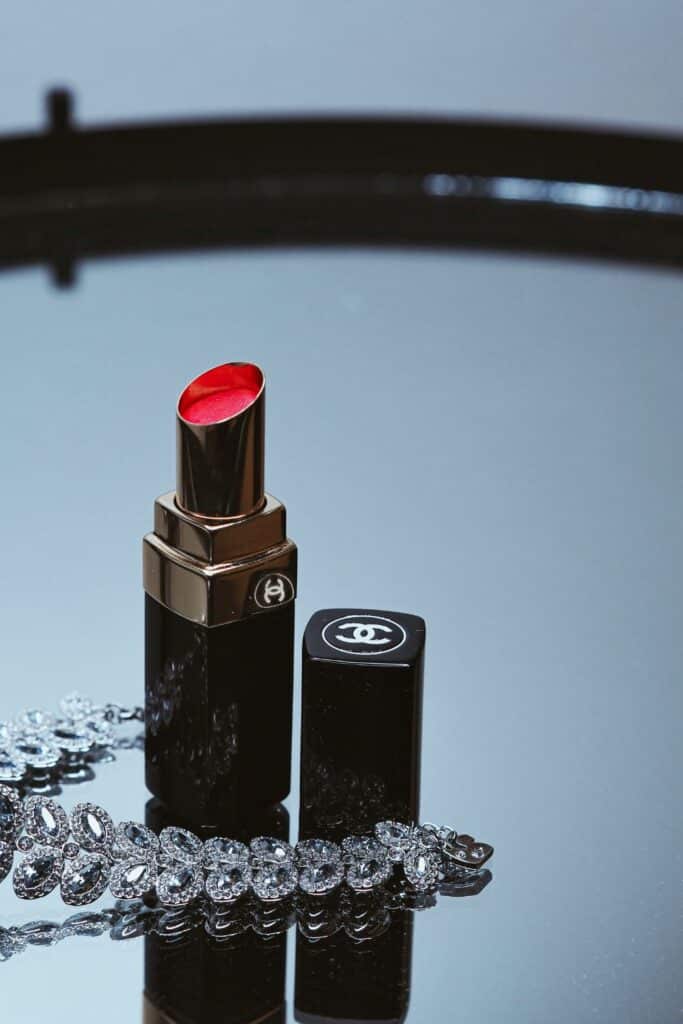 A luxurious red Chanel lipstick stands next to its sleek black case on a reflective surface, accompanied by a sparkling rhinestone bracelet—icons of Parisian chic beauty.