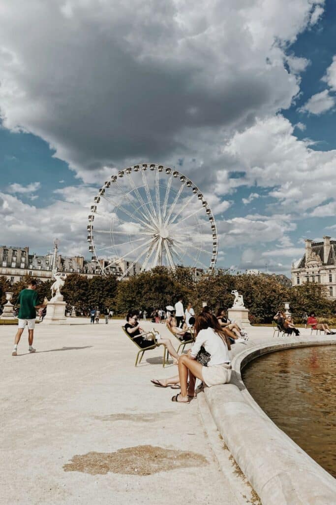 People relax in the Tuileries Garden under a dramatic sky, with a large Ferris wheel in the background. Parisian locals and tourists blend in with refined summer outfits, steering away from overly sporty looks or loud prints.