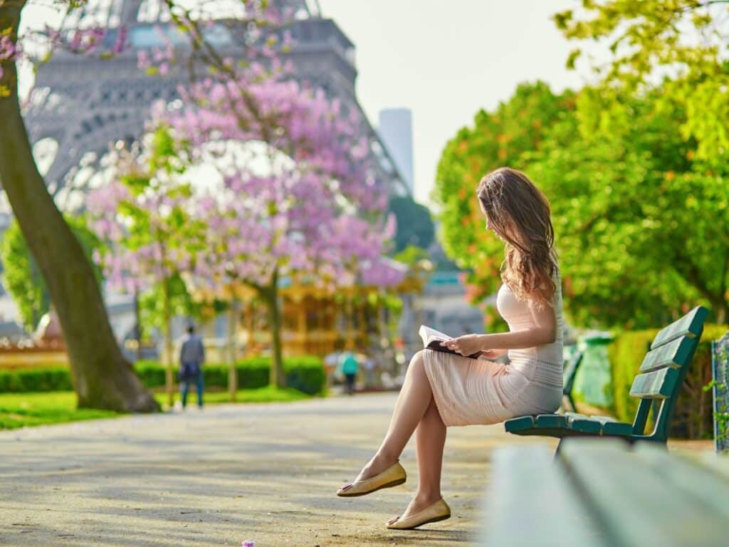 A woman reads on a park bench near the Eiffel Tower surrounded by blooming trees, wearing a blush pink knit dress and nude flats. Her soft, coordinated look reflects thoughtful styling and avoids short shorts or athletic gear.