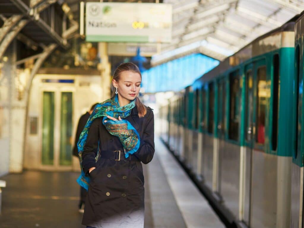 A woman walks along a train platform in a belted black coat and a bright blue patterned silk scarf, merging travel ease with Parisian chic sophistication.