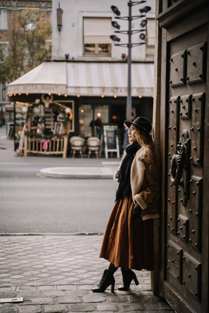 A woman dressed in a rust-colored skirt, heeled boots, a shearling jacket, and a wide-brim hat leans against a doorway in a cozy Parisian street scene. This layered, textured look avoids the faux pas of skipping accessories or wearing overly revealing clothing.