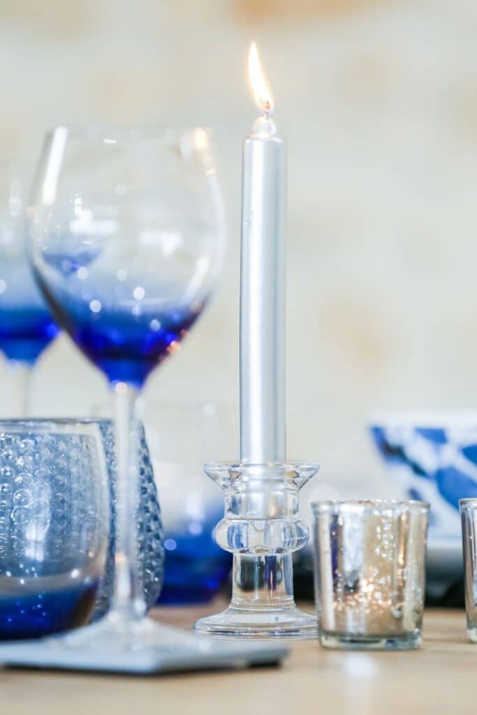 A lit silver taper candle surrounded by shimmering votive holders and ombre blue glassware sets a warm, inviting atmosphere for a French Country style table.