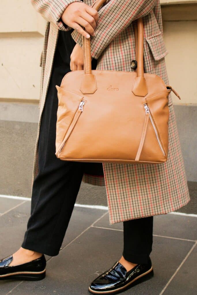 A close-up of a structured tan leather handbag with silver zippers, carried by a woman in a plaid coat and black pants—an essential piece of the Parisian chic wardrobe.